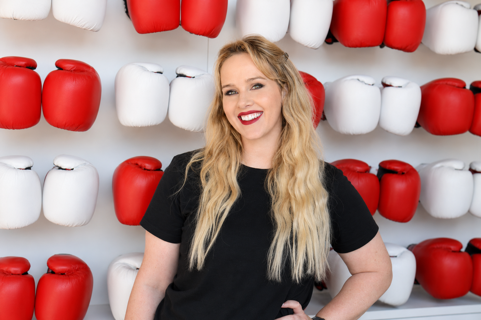 A woman with long blonde hair and red lipstick standing in front of a wall of red and white boxing gloves, smiling at the camera.
