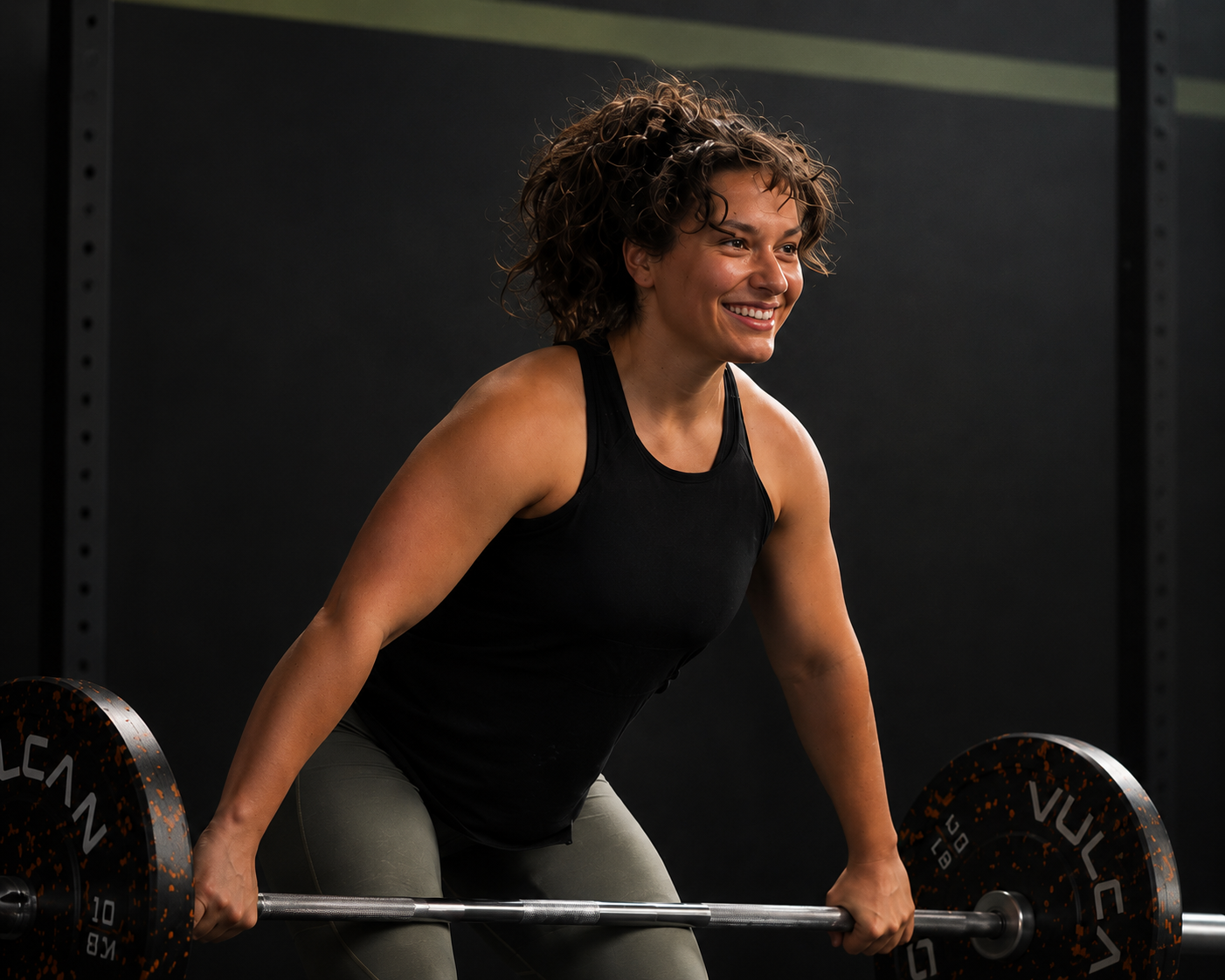 A woman with curly hair smiling while lifting a barbell in a gym setting.