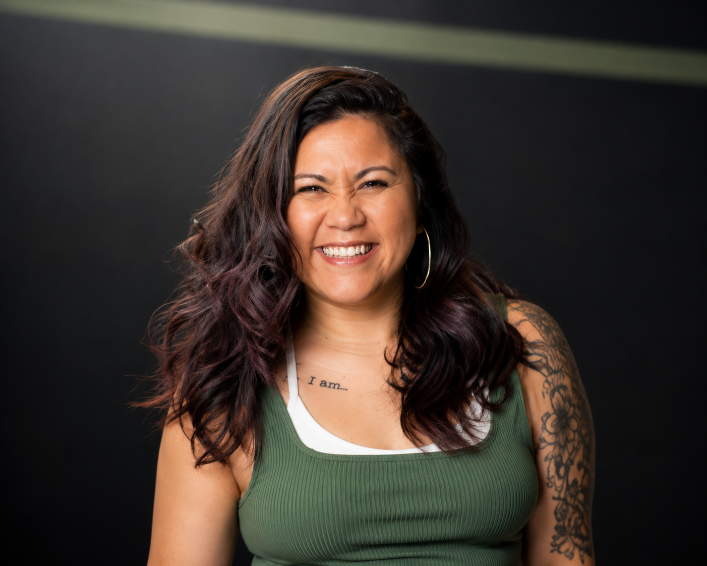 A woman with long dark hair and tattoos, winking and smiling in front of a dark background.