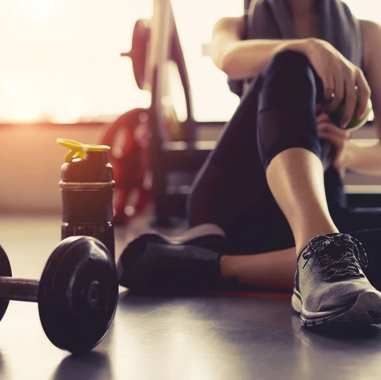 A person sitting on the gym floor with a water bottle, dumbbell, and fitness gear nearby, preparing for or resting from a workout.
