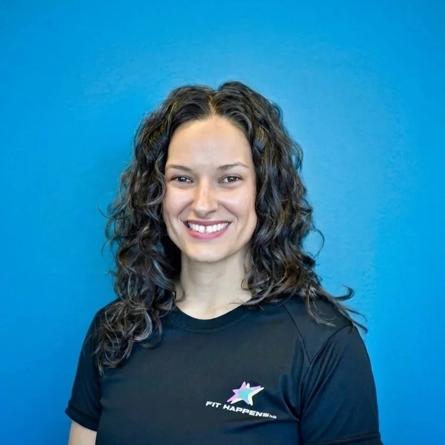 A woman with curly hair smiling while lifting a barbell in a gym setting.