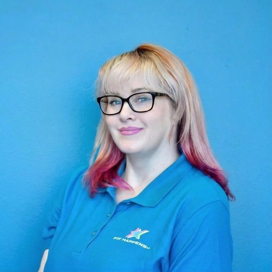 A woman with blonde hair, glasses, and a black top sitting at a reception desk with a sign that reads 'Welcome, we're glad you're here!' behind her is a black wall with the words 'Stronger Together, Fit Happen' and a shelf with black items, a window, and a potted plant.
