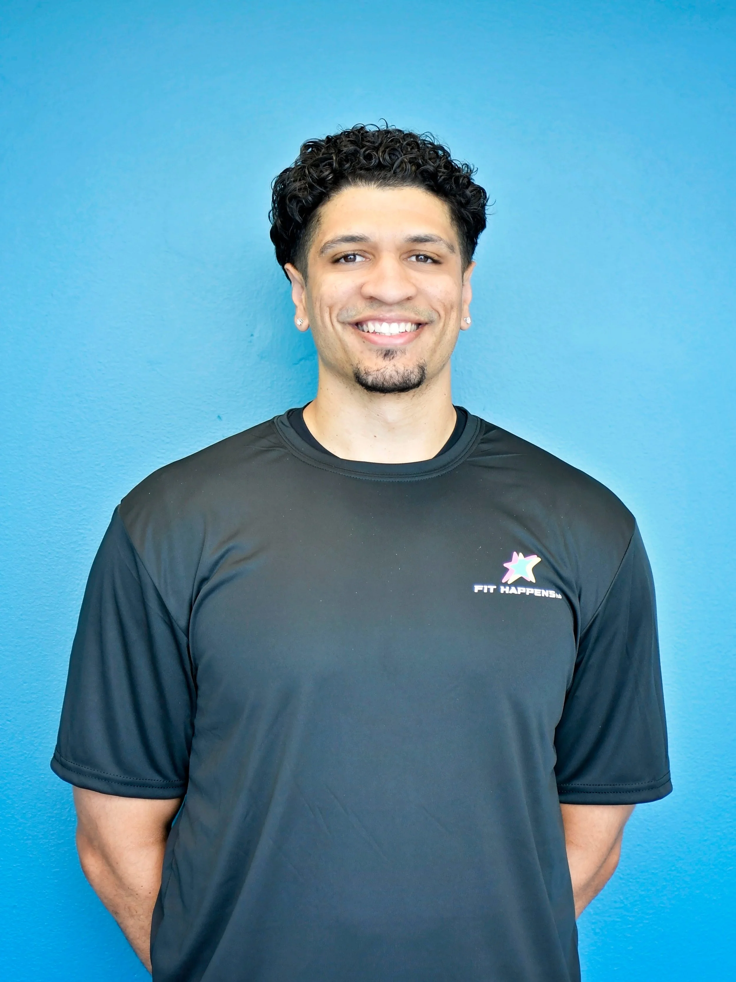 A smiling man with curly dark hair, earrings, and a goatee wearing a black long-sleeve shirt, standing against a dark brick wall background.