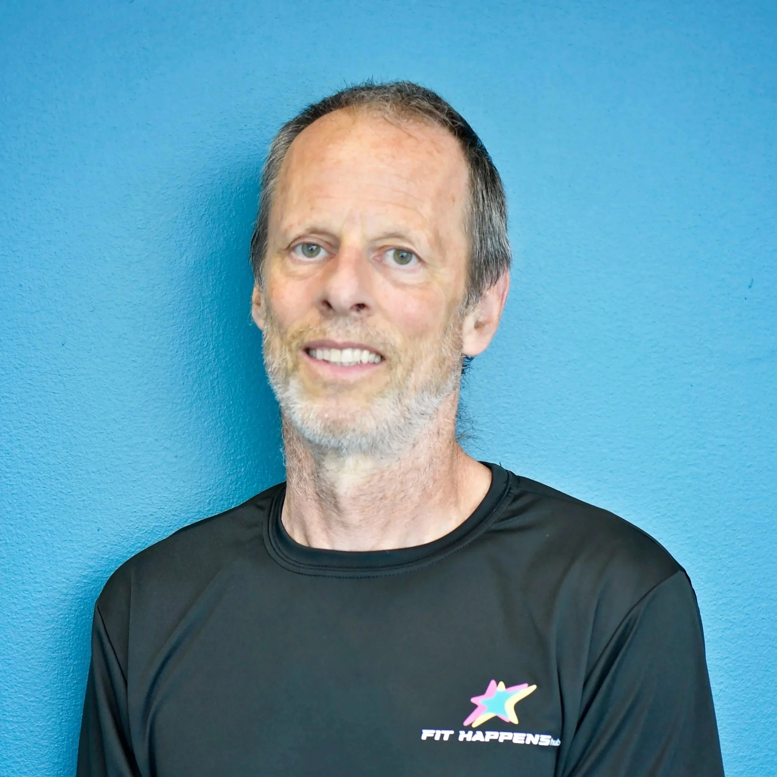 A middle-aged man with a beard and short hair smiling in a gym with black brick walls and dumbbells in the background.