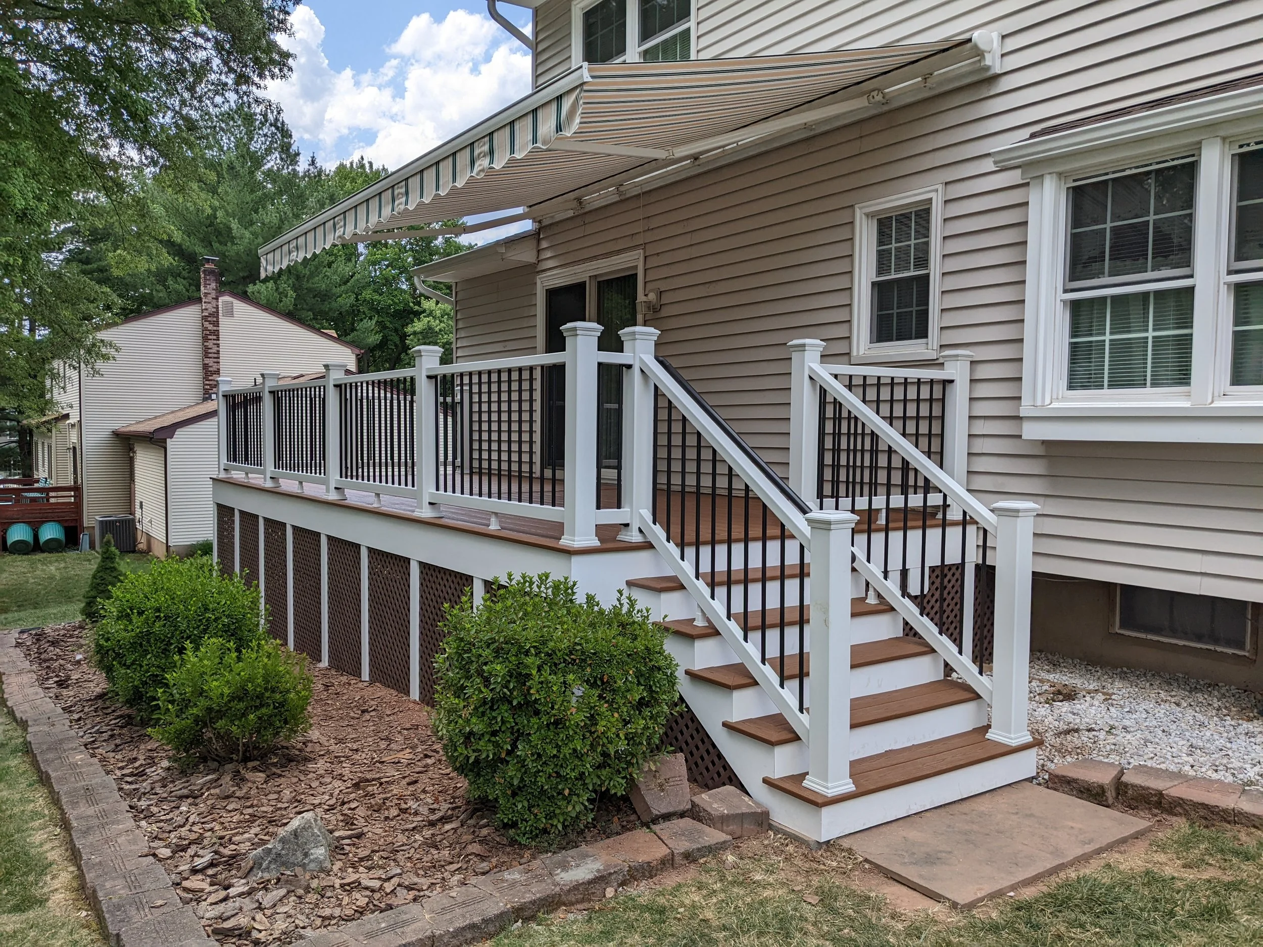 A backyard patio with a house, stairs, and a striped retractable awning. There are trees and neighboring houses visible in the background.