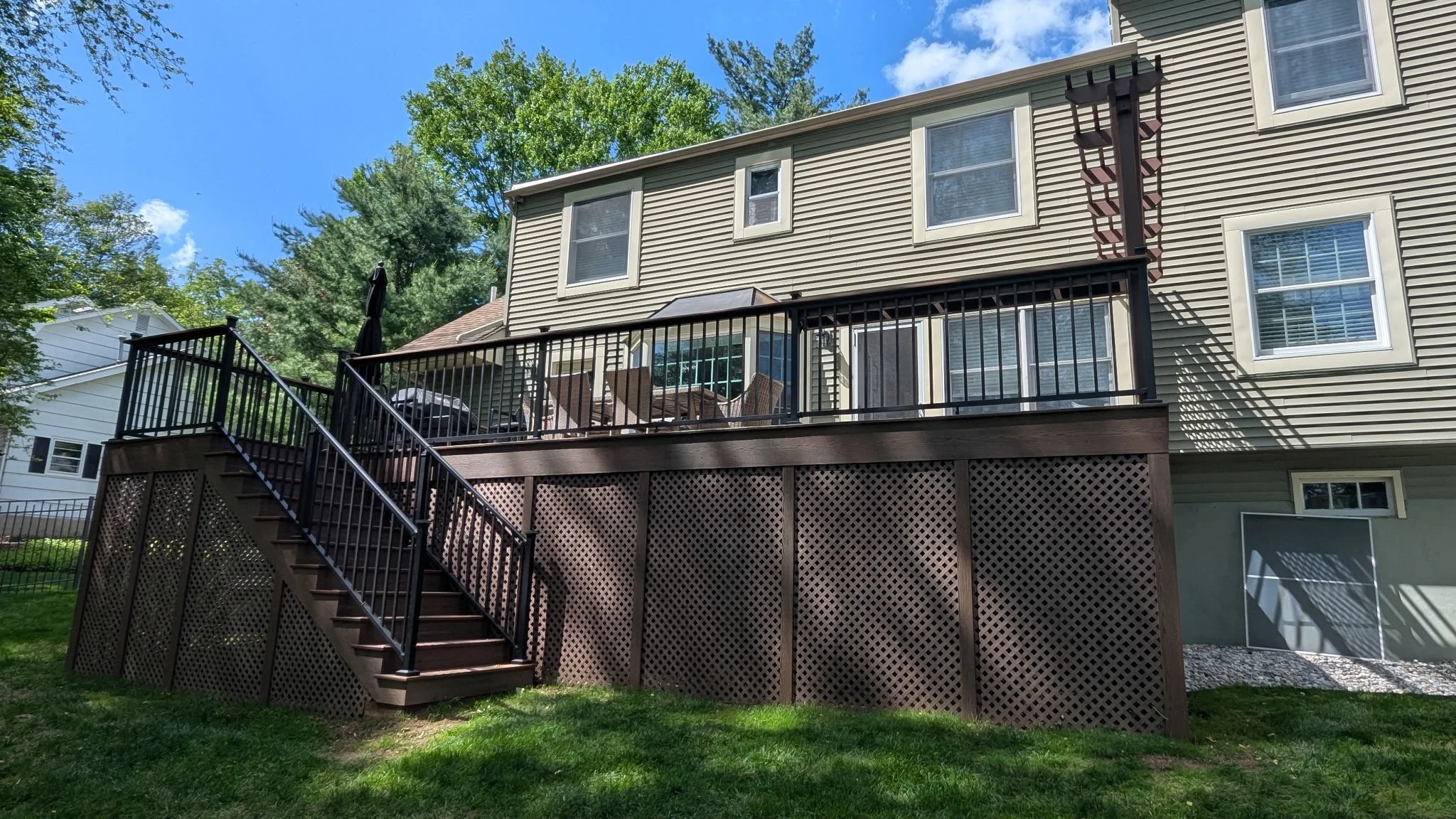 Backyard view of a two-story house with a wooden deck, black railing, set of stairs, and surrounding green lawn and trees under a partly cloudy sky.