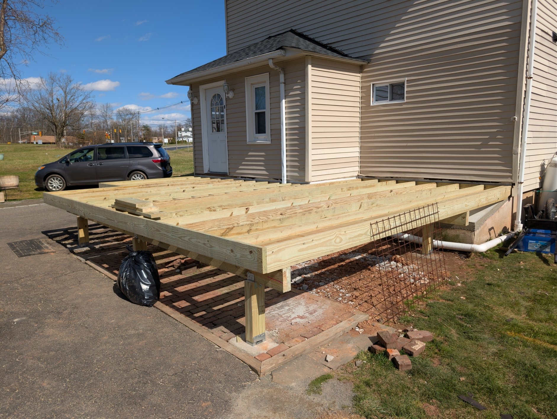 Construction site of a wooden deck attached to the back of a beige house. The deck's support structure is being built with large wooden beams. A black garbage bag is on the ground in front of the deck. A gray minivan is parked nearby under a clear blue sky with some clouds.