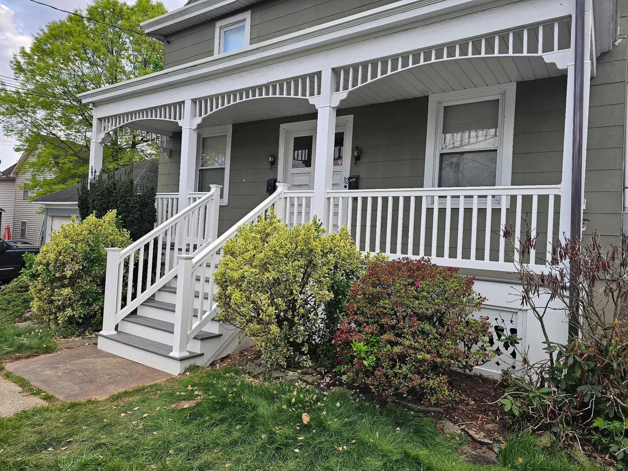 Front porch of a house with light green siding, white decorative trim, and a white staircase leading to the door. There are two front windows with blinds, and outdoor light fixtures near the door. Green bushes and plants are in the yard, and a large tree is visible in the background.