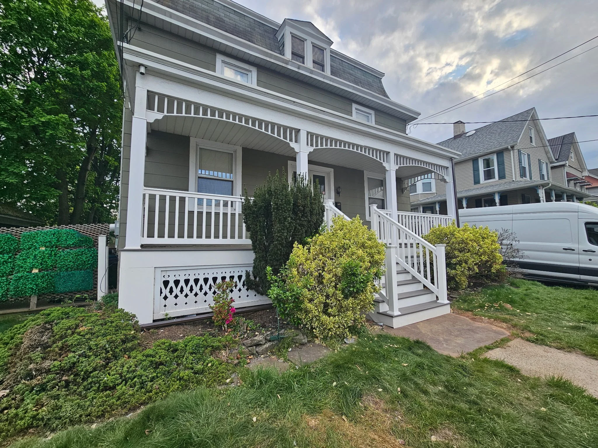 Front view of a gray house with white porch railings and stairs, green shrubs in the garden, neighboring houses, a white van parked on the street, and a partly cloudy sky.