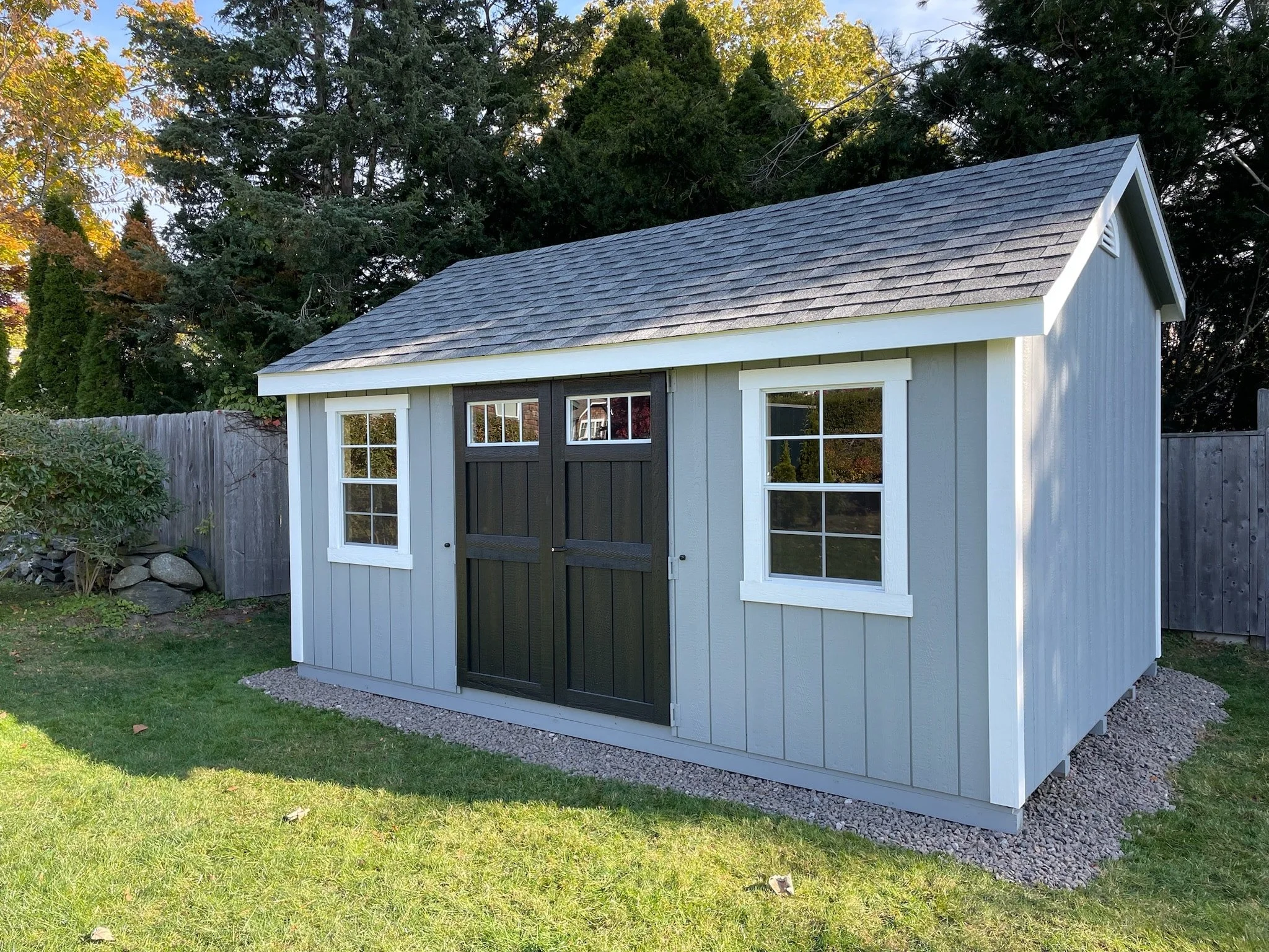 A small gray wooden shed with white window frames and black doors, situated in a backyard with a green lawn and a gravel base. Tall evergreen trees and some deciduous trees with autumn-colored leaves are in the background.