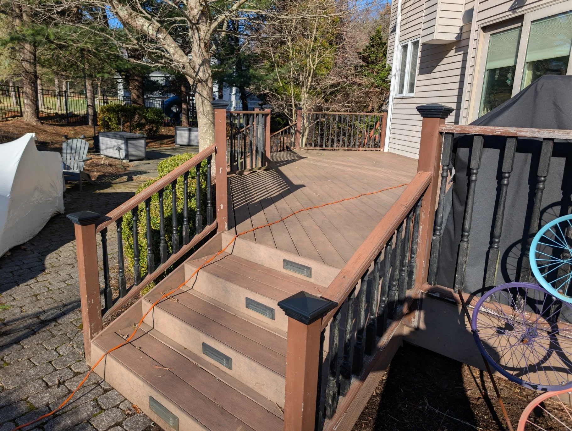 A wooden outdoor deck with stairs and railing, attached to a house with beige siding. Several trees and plants surround the area, and a black grill is partially covered on the right. There are some chairs, planters, and children's bicycles nearby.