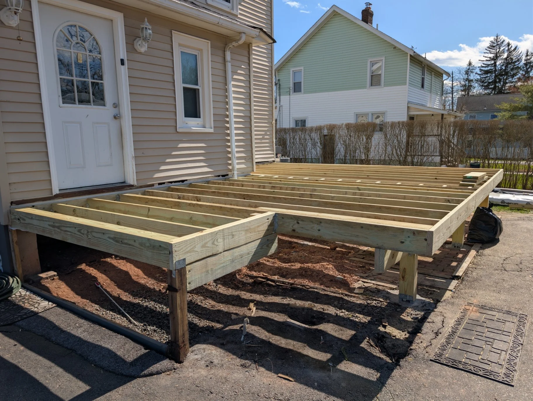 Construction site with a wooden deck frame in progress attached to the back of a house, with neighboring houses visible in the background under a partly cloudy sky.