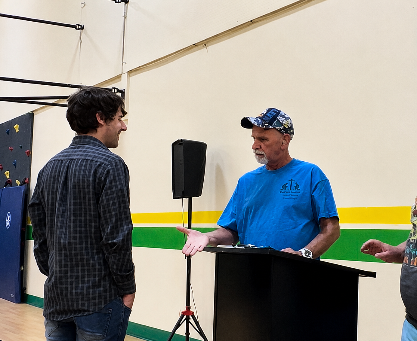 Ron McCoy, Independent candidate for WA State Senate 7th District, having a conversation indoors with a community member. Ron is facing the camera, wearing a blue t-shirt and baseball cap, with gray hair and a beard.