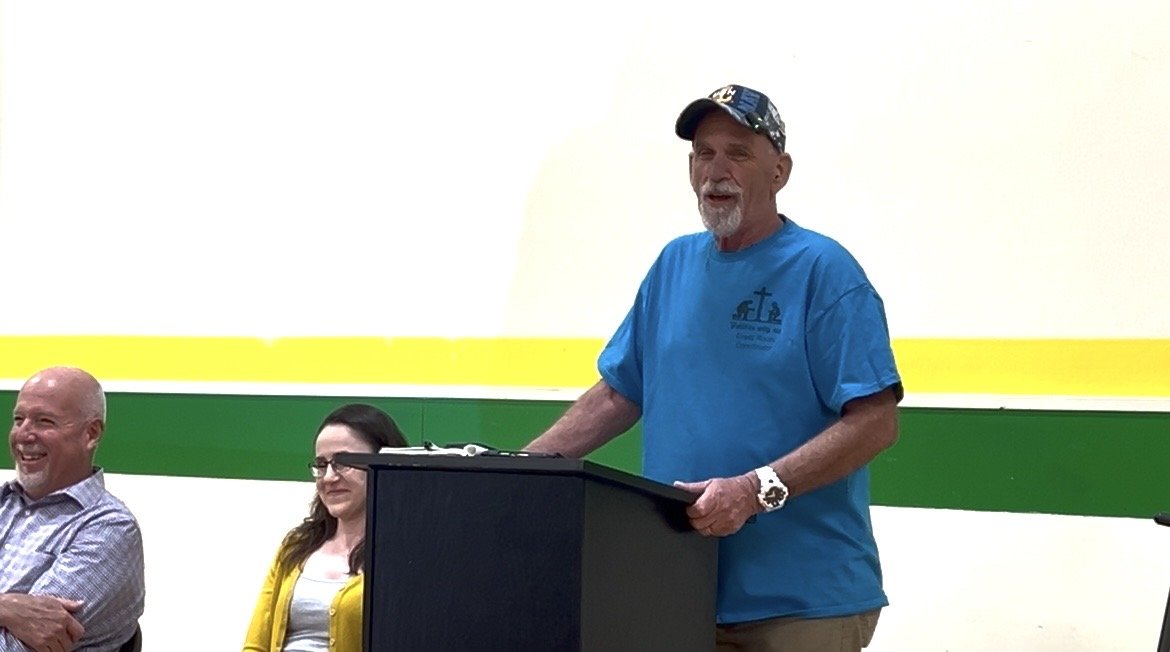 An older man with a gray beard and wearing a blue T-shirt and cap standing at a podium, speaking or giving a speech in a room with a white wall and a green and yellow stripe.