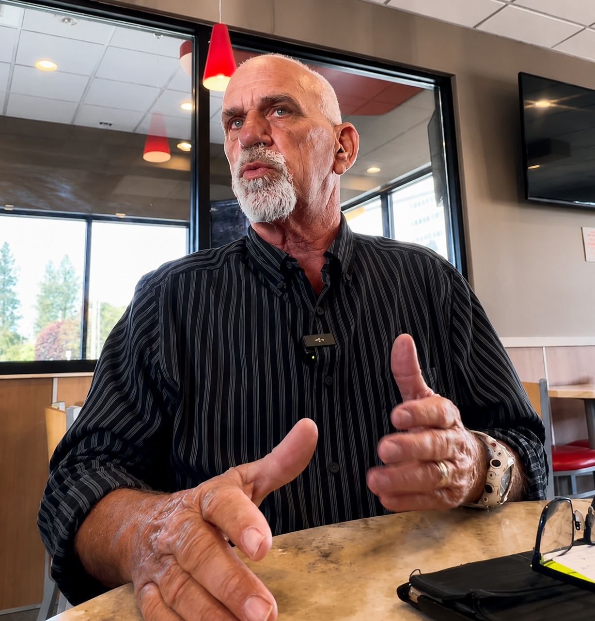 Ron McCoy, Independent candidate for WA State Senate 7th District, sitting at a table inside a restaurant. Ron has white hair and a beard and is wearing a black striped shirt, gesturing with his hands during a conversation.