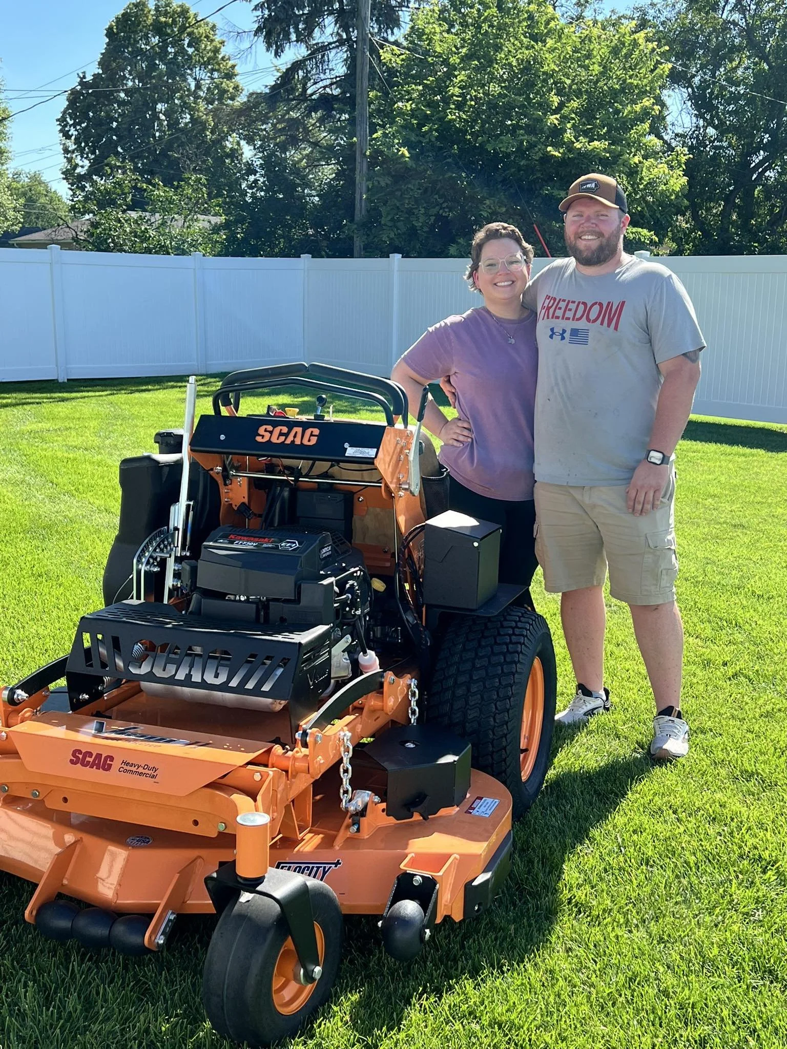 A couple stands behind a commercial turf mower in a backyard with green grass and a white fence, trees, and a blue sky in the background.