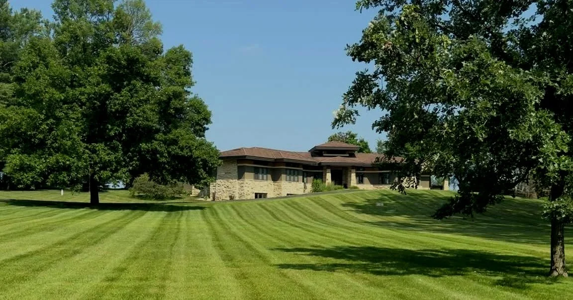 Large house with a stone and stucco exterior, surrounded by a vast, well-maintained grassy lawn with evenly trimmed stripes and mature trees on a sunny day.