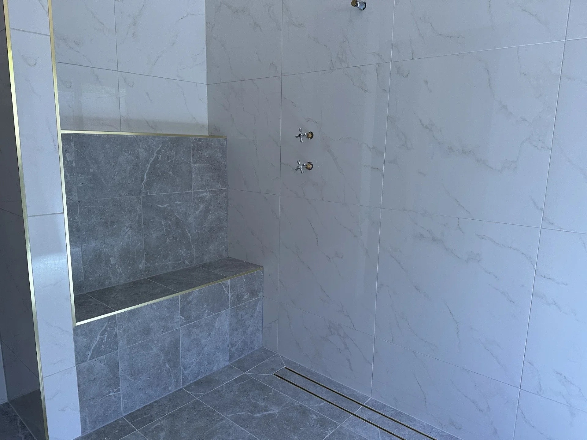 A modern shower with a built-in bench, gray marble tiles, and a metal showerhead on a white marble-tiled wall.