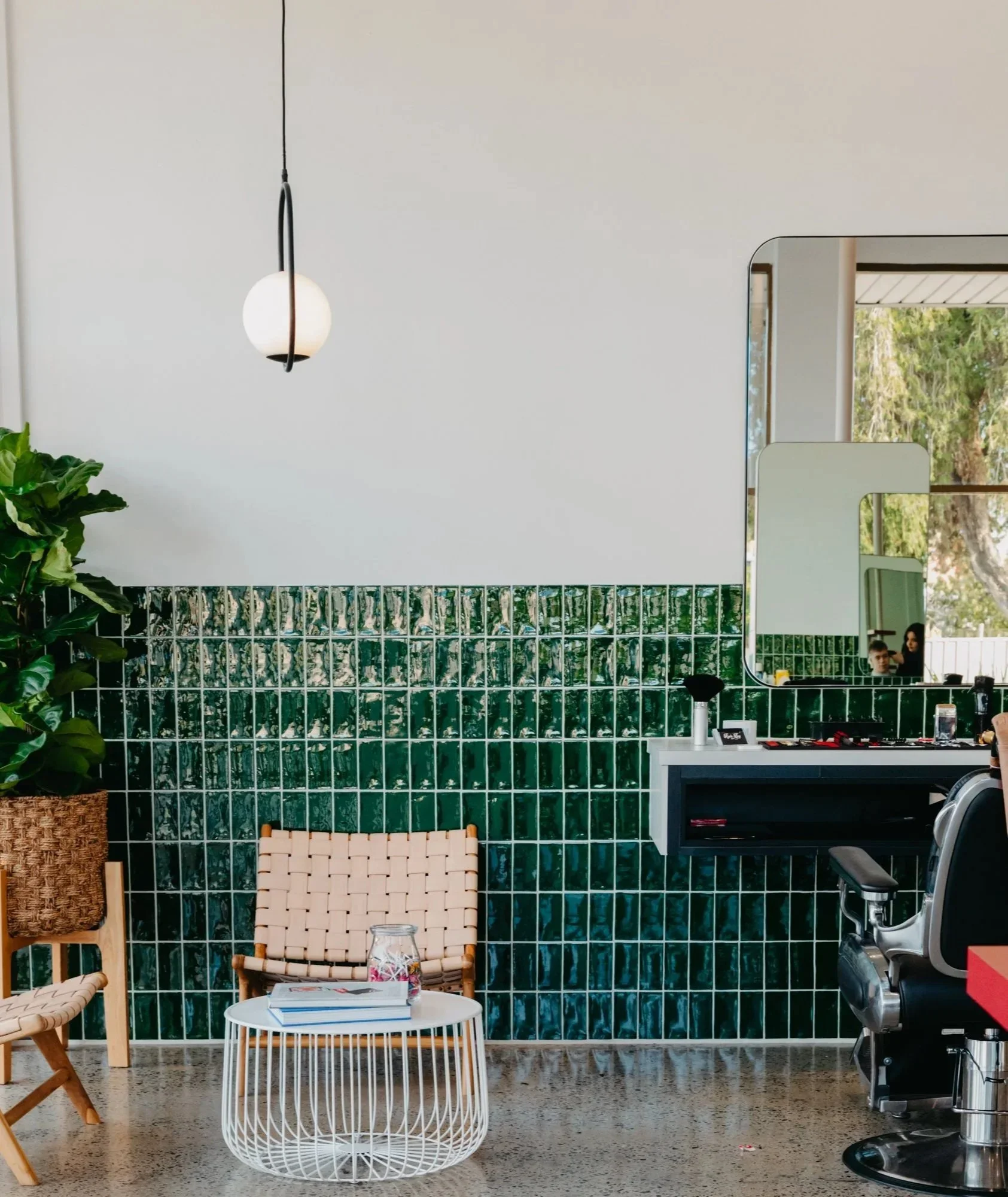Interior of a modern room with a hanging white pendant lamp, a green tiled wall, and a window showing trees outside. There is a white round coffee table with magazines, a wooden chair, a black chair, a barber's station with a mirror, and a barber's chair.