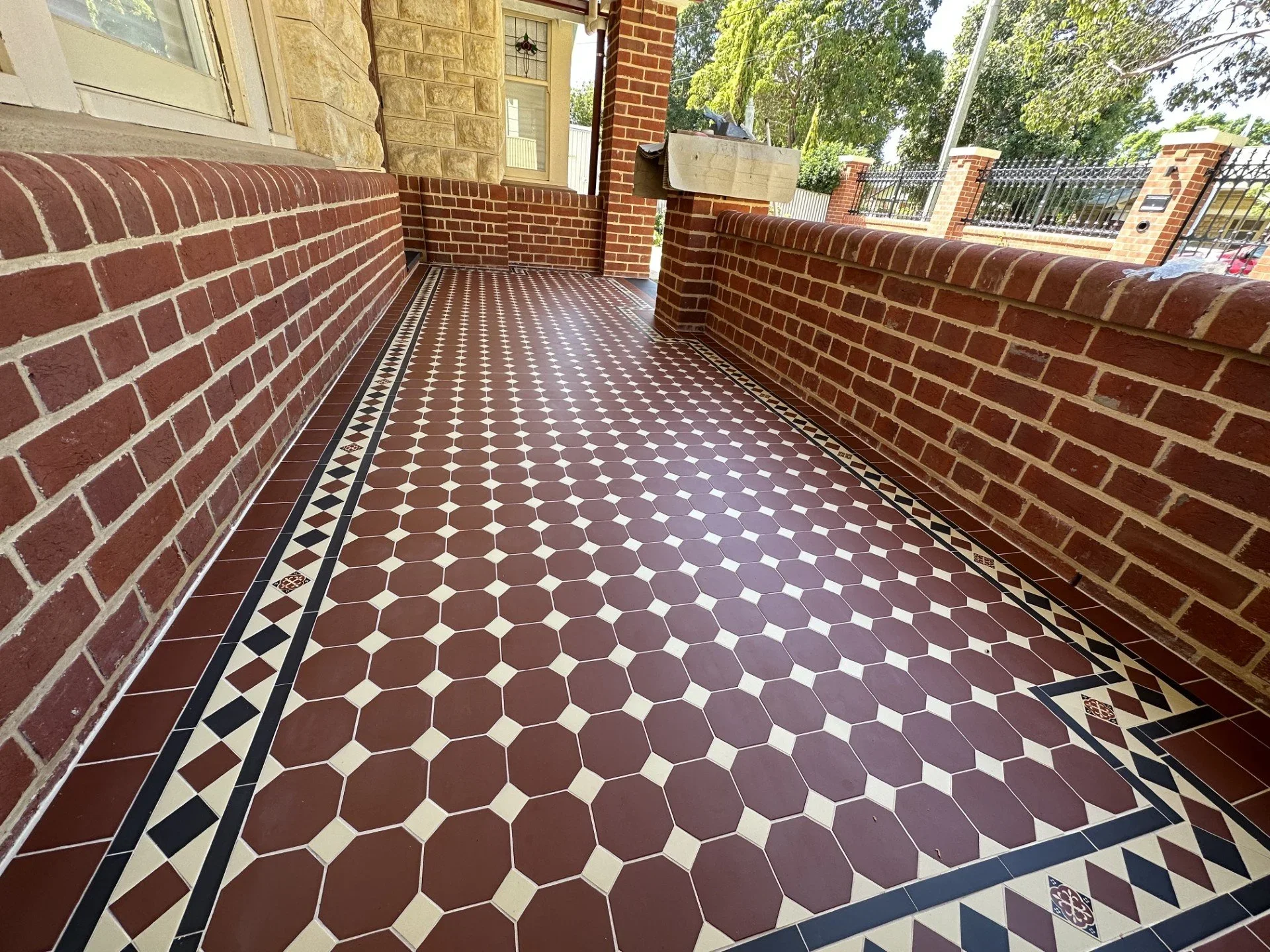 A porch with a decorative tiled floor featuring red hexagonal tiles with a black and white border. The porch is enclosed by red brick walls and a brick pillar, with green trees and a metal fence visible beyond.
