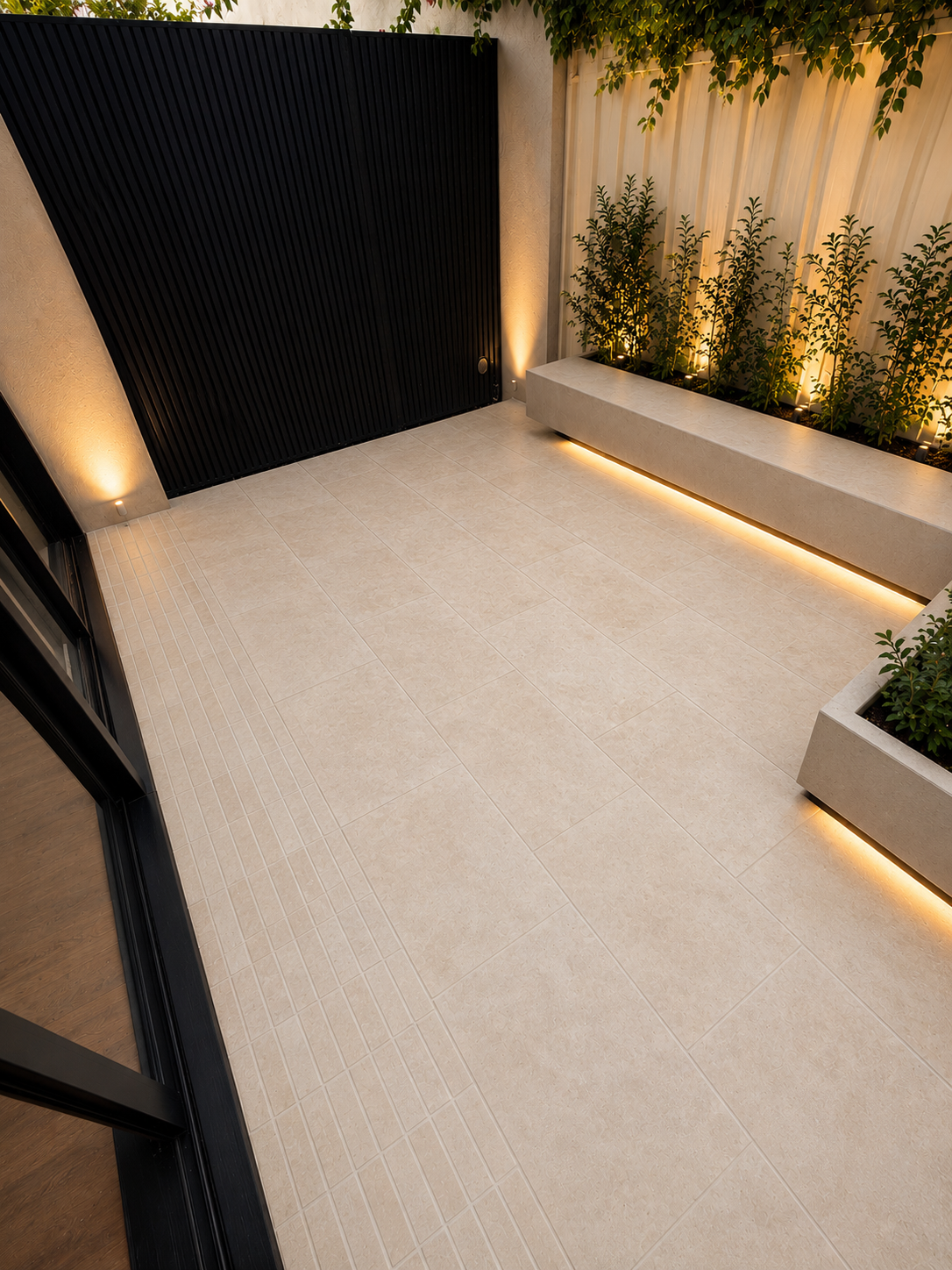 A modern outdoor patio area with beige tiled flooring, a black vertical slatted gate, and a beige concrete planter with green plants and accent lighting.