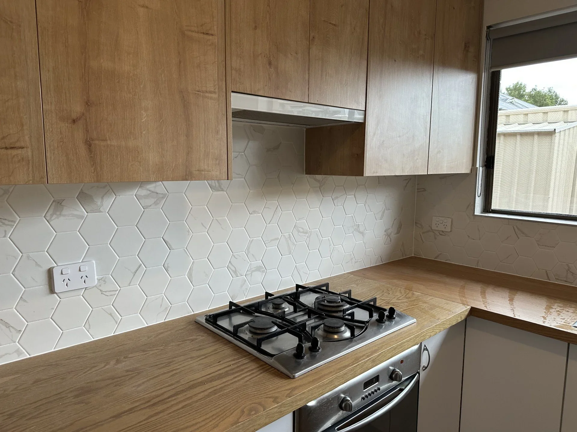 Kitchen with wooden cabinets, white hexagonal marble backsplash, gas stove on wooden countertop, and window with blinds.