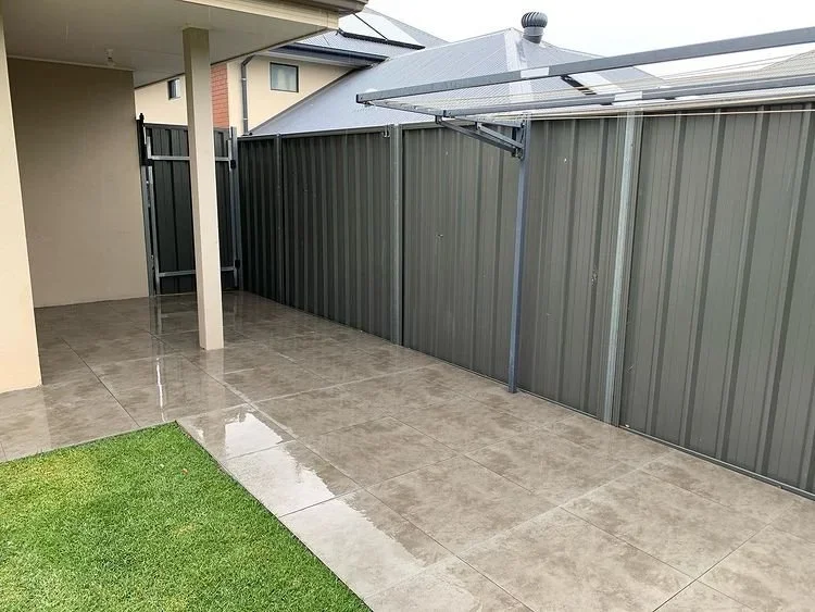 Backyard with wet concrete patio, small patch of artificial grass, gray metal fence, and a metal clothesline under a covered patio area.