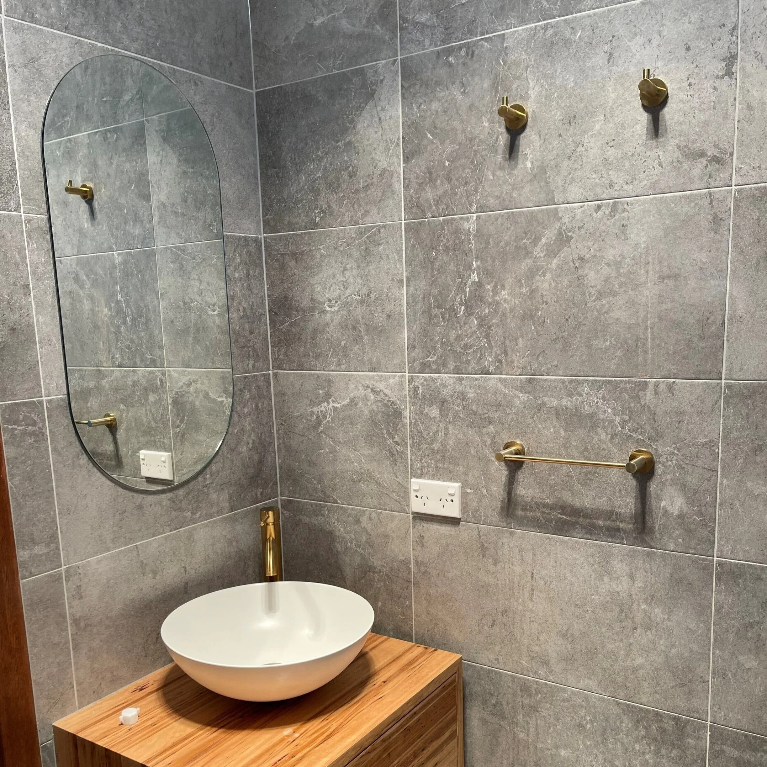 Gray tiled bathroom wall with a large oval mirror, two gold hooks, a wooden vanity with a white vessel sink, a gold faucet, and a power outlet.
