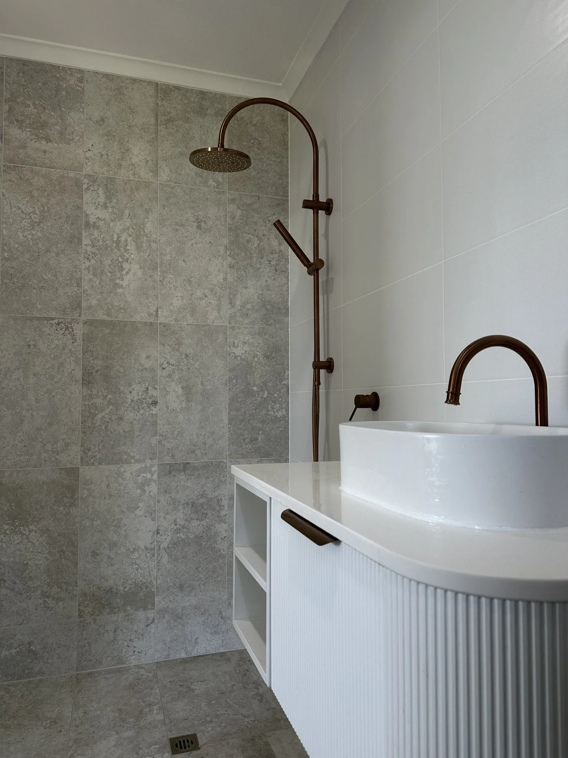 A shower area with a copper-colored rain showerhead and a handheld shower, gray tiled wall, white ceiling, and a white vanity with a rounded white sink.