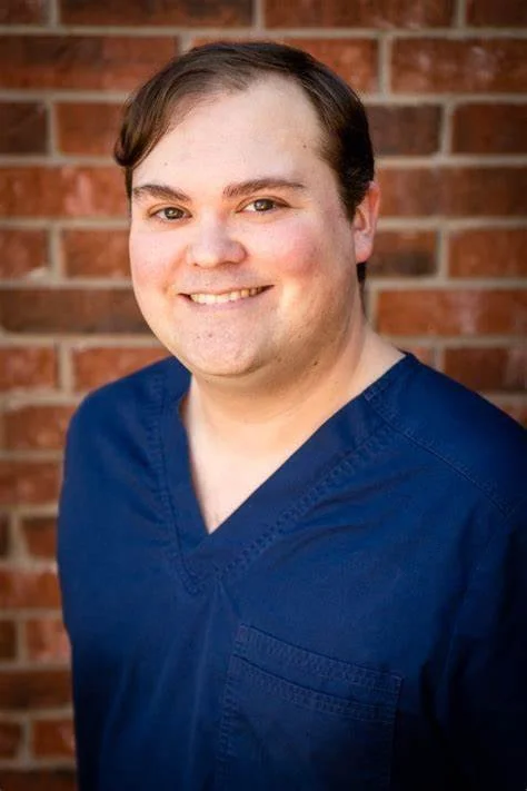 A man with short brown hair and fair skin wearing blue medical scrubs, standing outdoors in front of a brick wall, smiling at the camera.