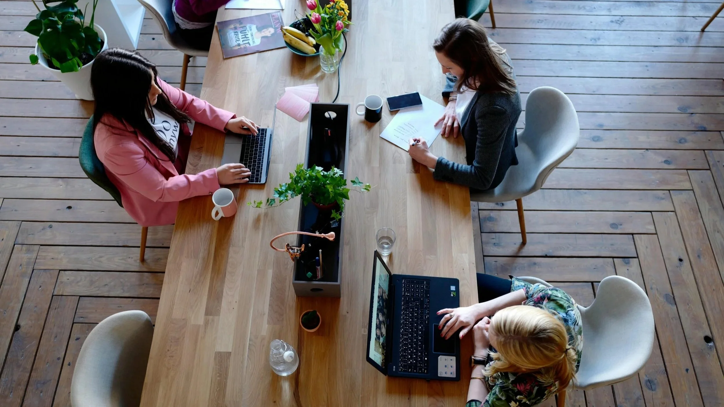 A top-down view of four women sitting at a wooden table working on AI SEO on laptops and writing SEO strategy notes. The table has plants, cups, a book, and a bowl of bananas with pink tulips. The floor is wooden.