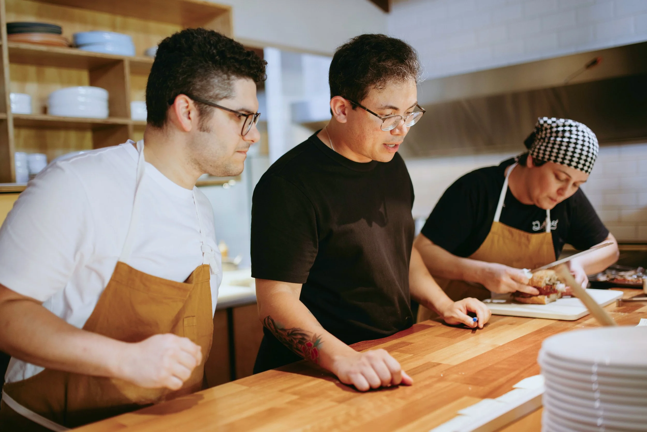 A manager and two coworkers preparing food together in a commercial kitchen., wearing aprons, preparing food at a wooden counter.
