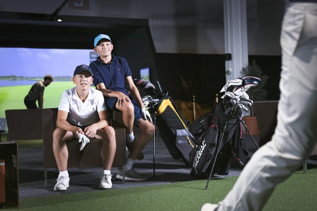 Two young boys at an indoor golf simulator, with golf bags and clubs nearby, one sitting on a bench and the other standing, while a golfer prepares to swing.