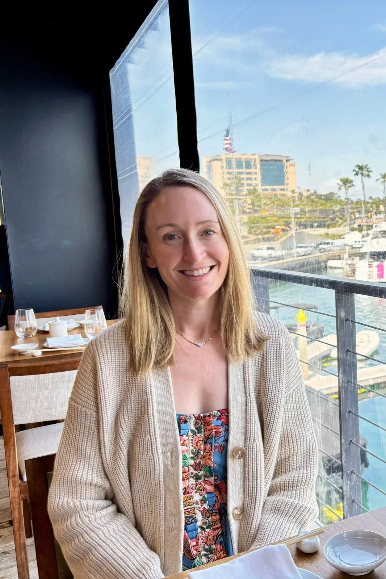 Katie Buzas, writer of The Layer Beneath, smiling and seated at a restaurant table with a marina view in the background.