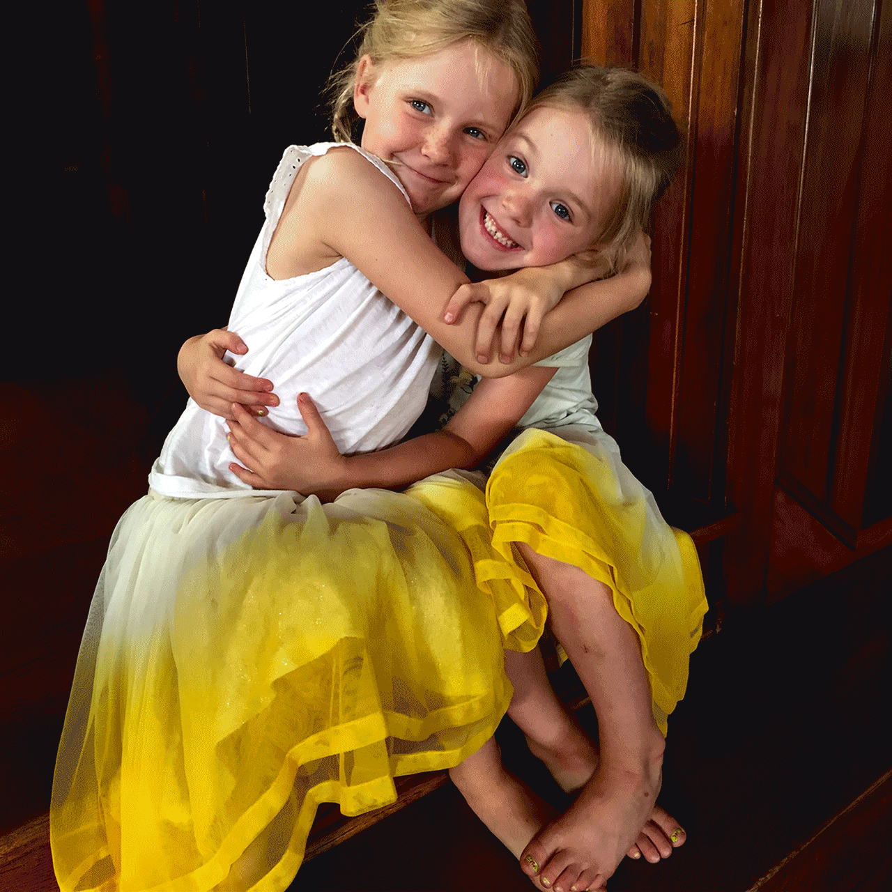 Two young girls hugging and smiling while sitting on a wooden bench, wearing white tops and yellow skirts, in a room with dark wooden walls.