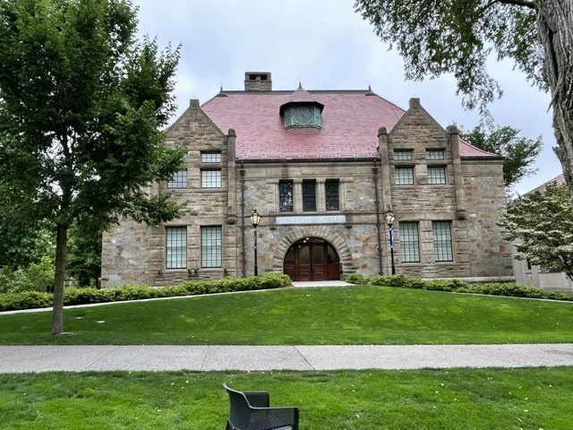 Historic stone building with a pink roof, situated on a well-maintained grassy area with trees and a sidewalk in the foreground.