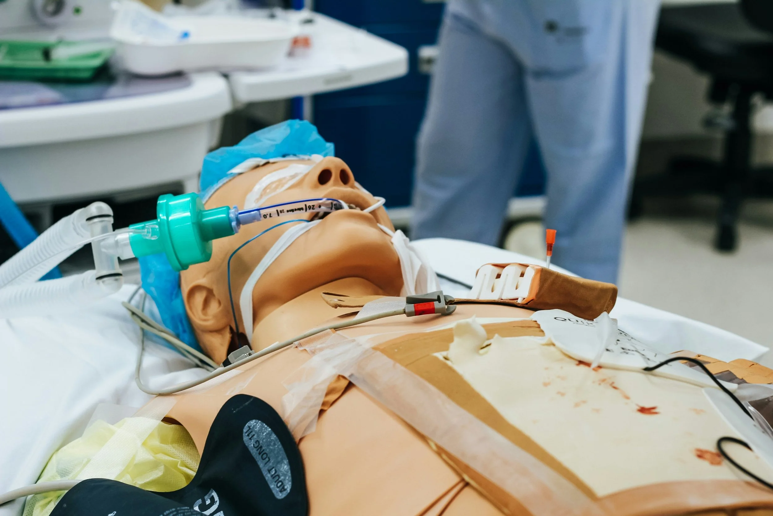 Medical simulation mannequin lying on hospital bed with artificial blood stains, connected to medical equipment including a breathing mask, monitoring wires, and IV lines, in a clinical setting.