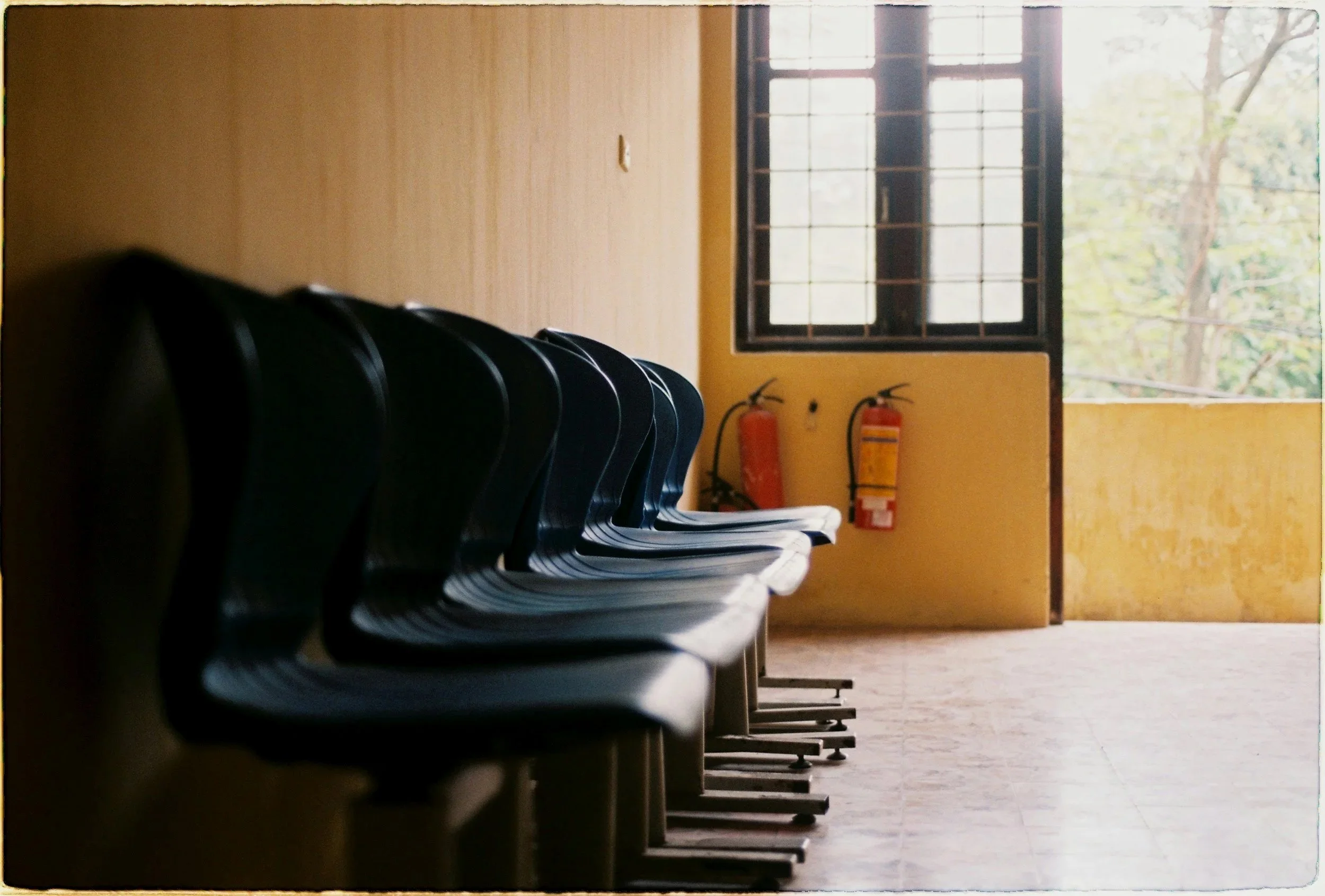 Empty rows of blue chairs in a room with a yellow wall, a window, and fire extinguishers.