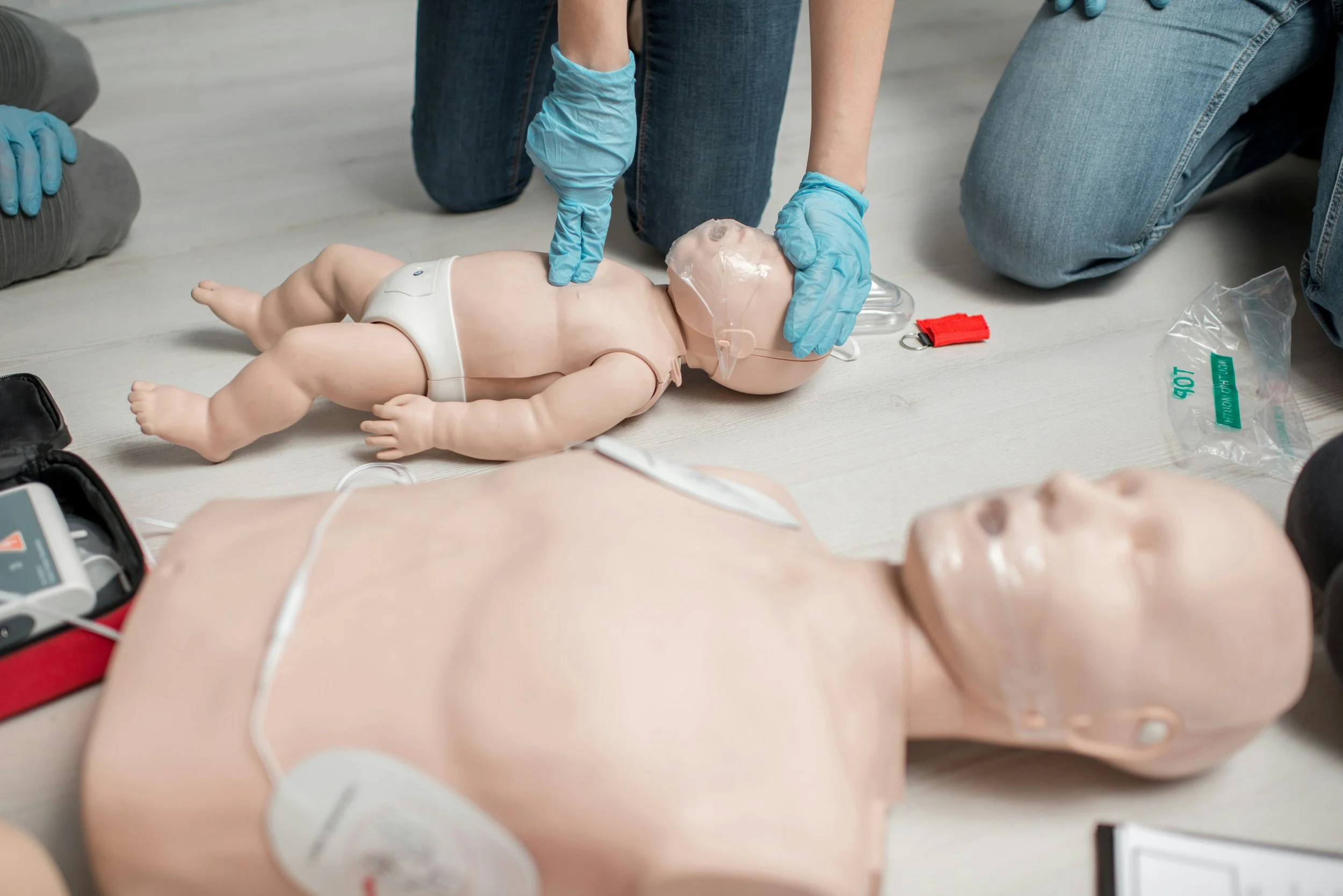 A medical training scene with a baby doll on the floor, a person wearing blue gloves performing CPR, with medical equipment nearby.