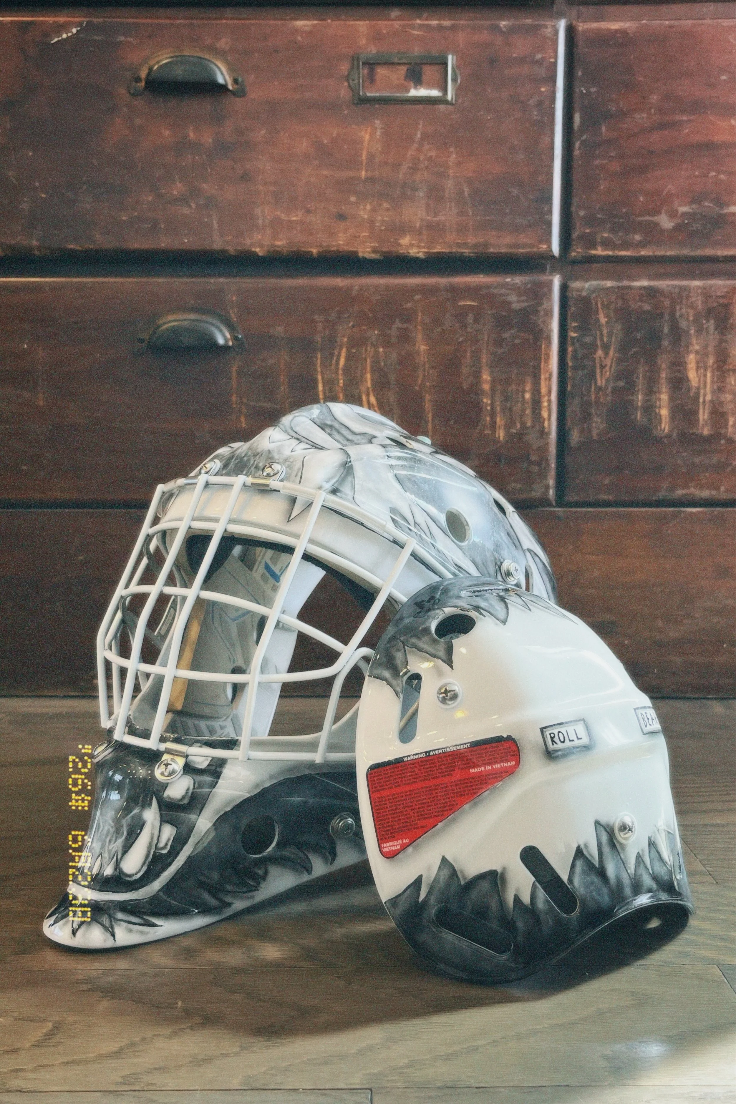 A hockey helmet with a painted aggressive shark face design, placed on a wooden surface in front of a dark wooden cabinet