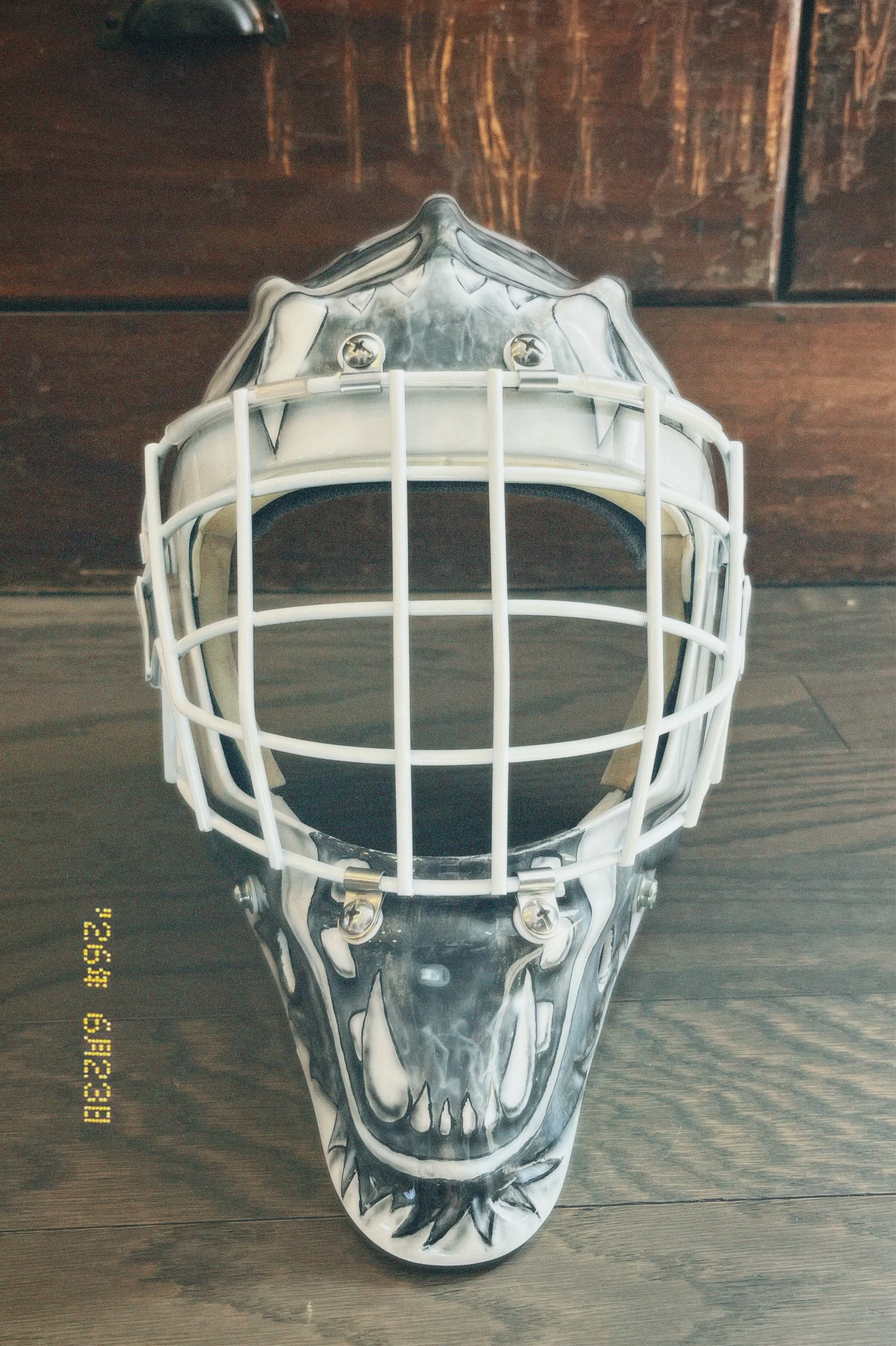 A white hockey goalie mask with a black and gray skull face design, placed on a dark wooden surface.
