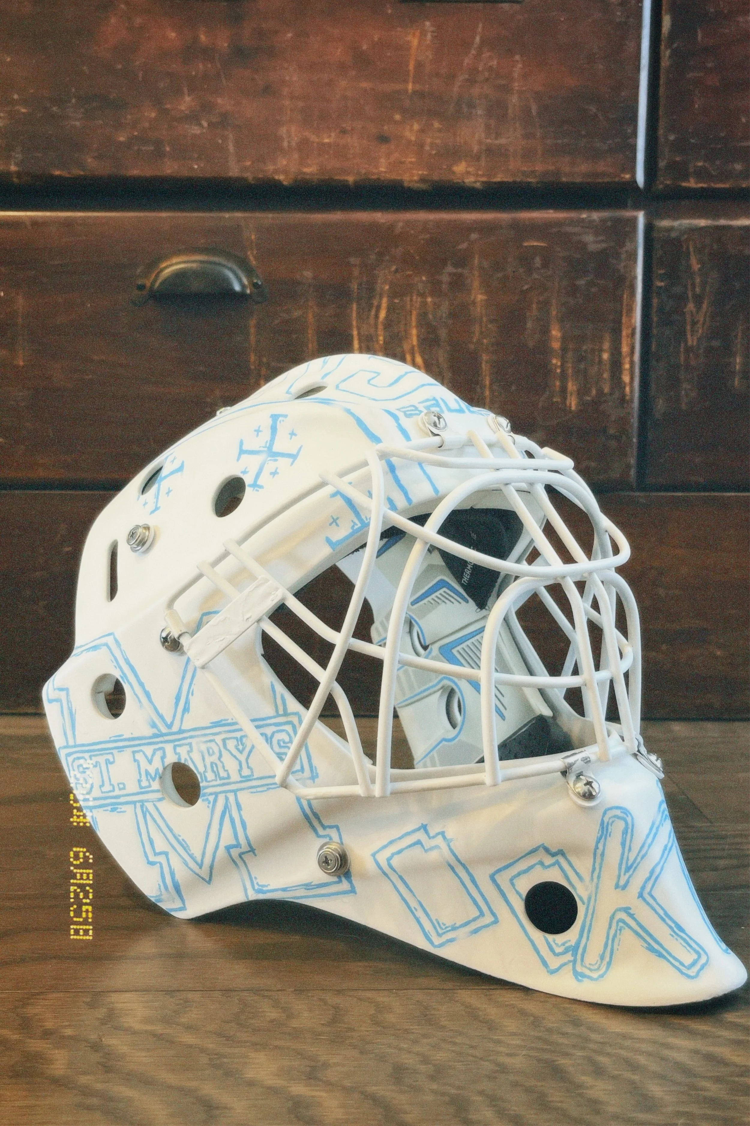 A white ice hockey goalie mask with blue designs resting on a wooden floor in front of a dark wooden dresser.