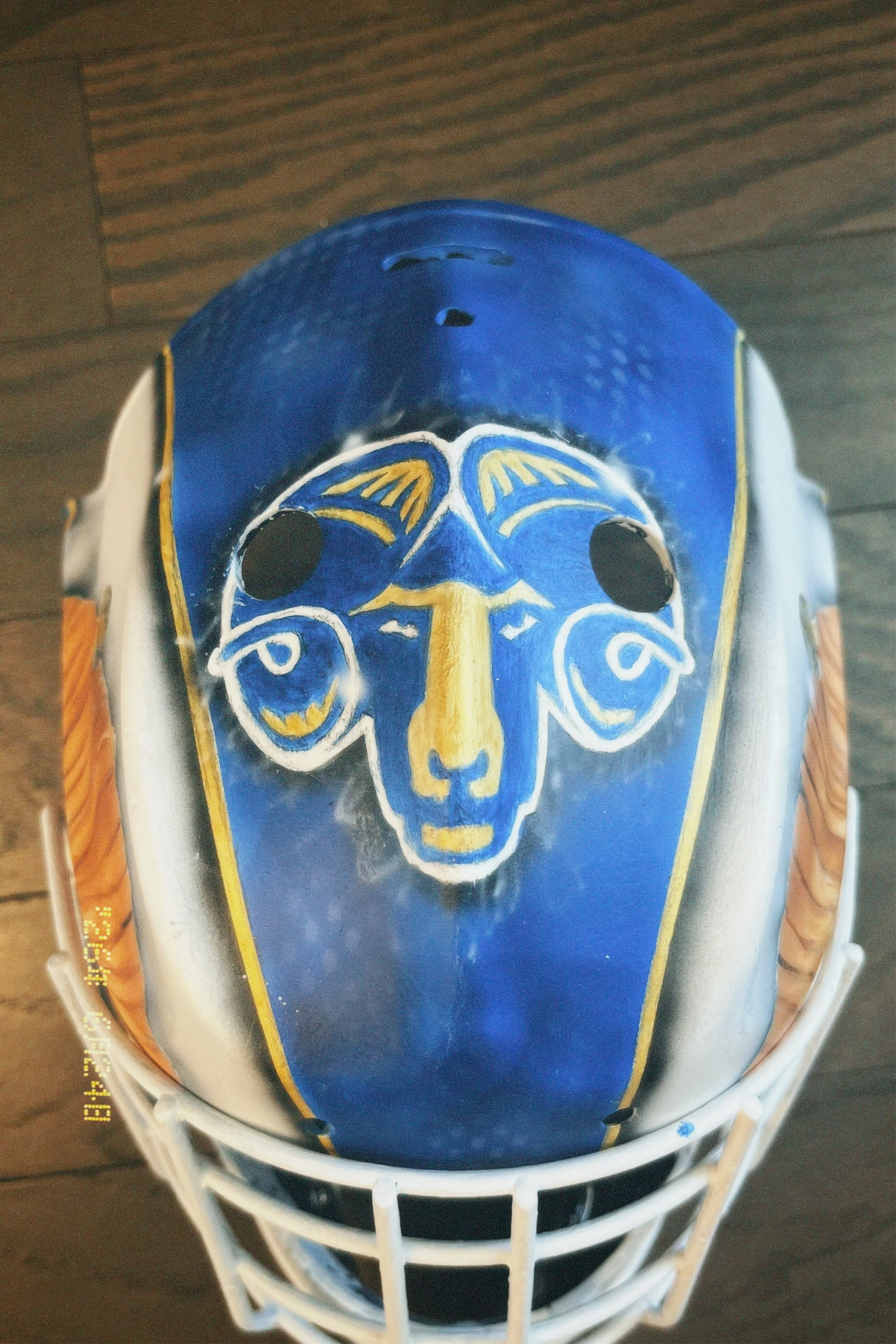 Close-up of a hockey goalie mask featuring a blue face with yellow and white accents, black eye holes, and a stylized face design, placed inside a hockey goal net on a wooden surface.