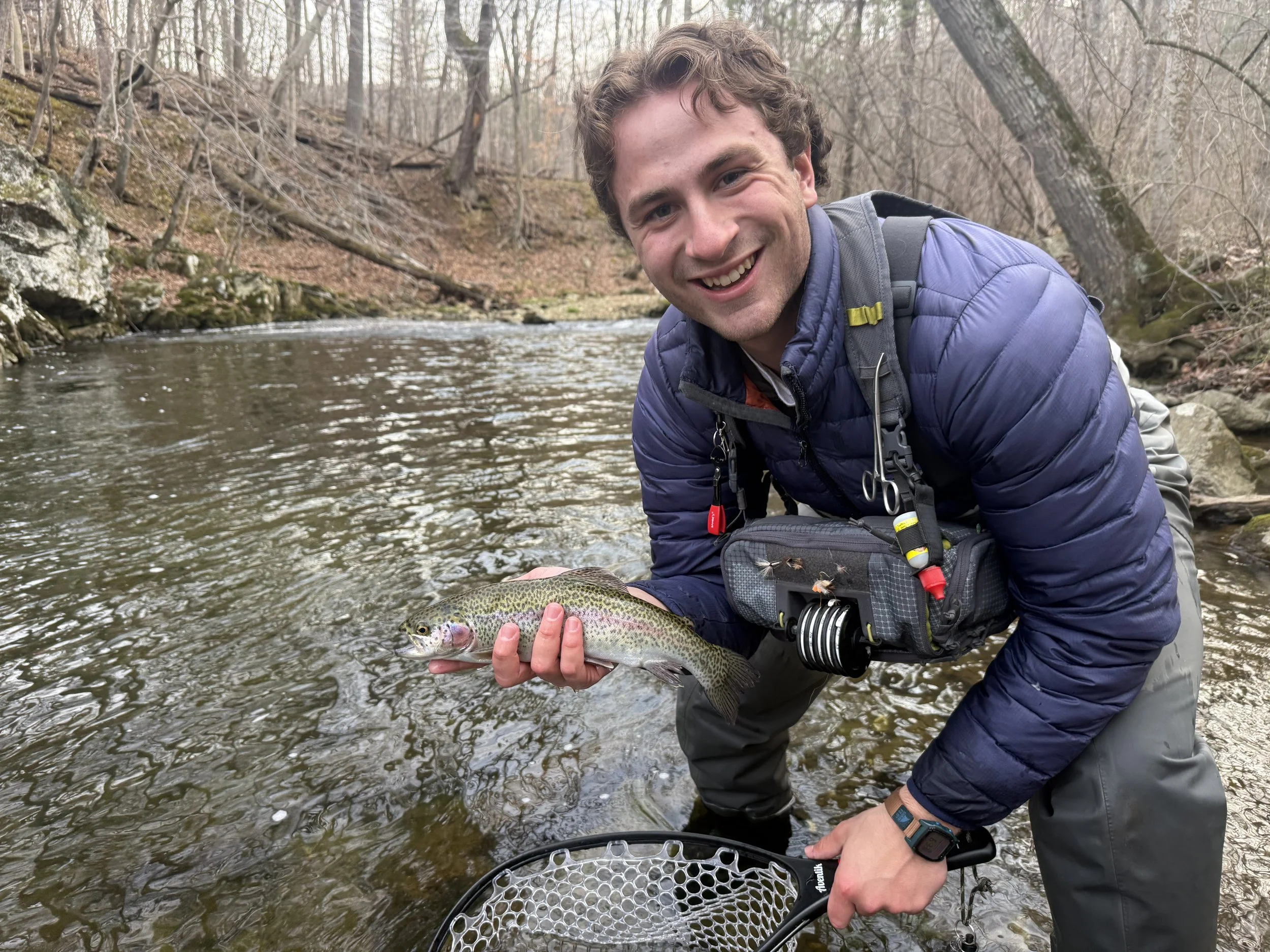 A smiling man with curly brown hair in outdoor clothing, holding a rainbow trout he caught while standing in a shallow river in a wooded area.