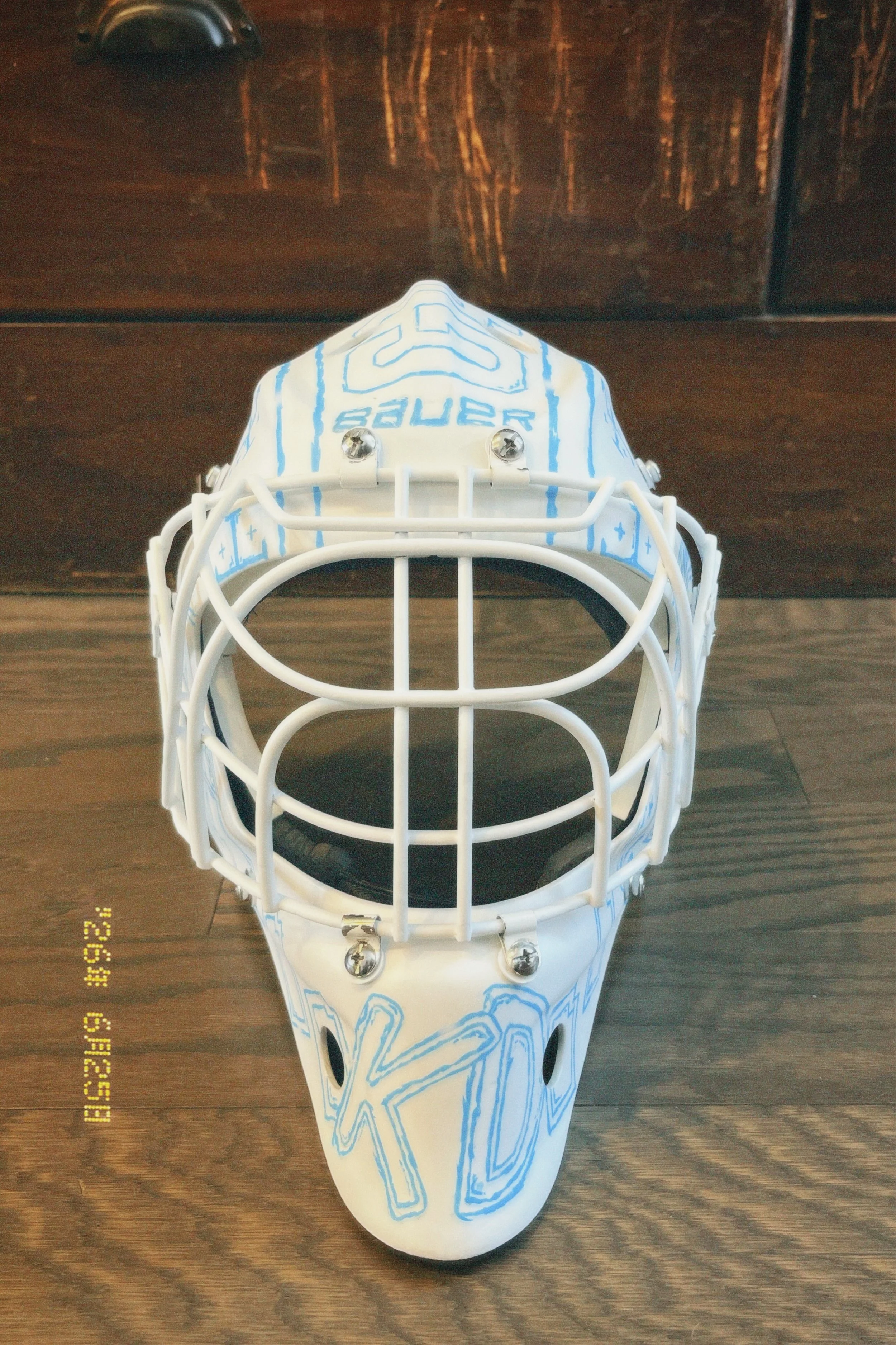 A white hockey goalie mask with blue designs on it, placed on a wooden floor.