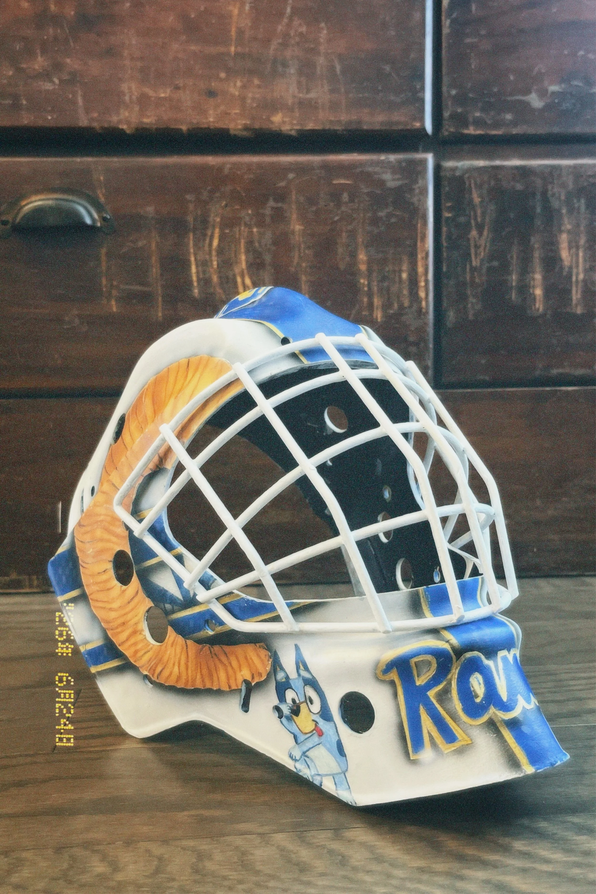 Ice hockey goalie mask with cartoon dog graphic and paw print, placed on a wooden surface with a dark wooden drawer in the background.