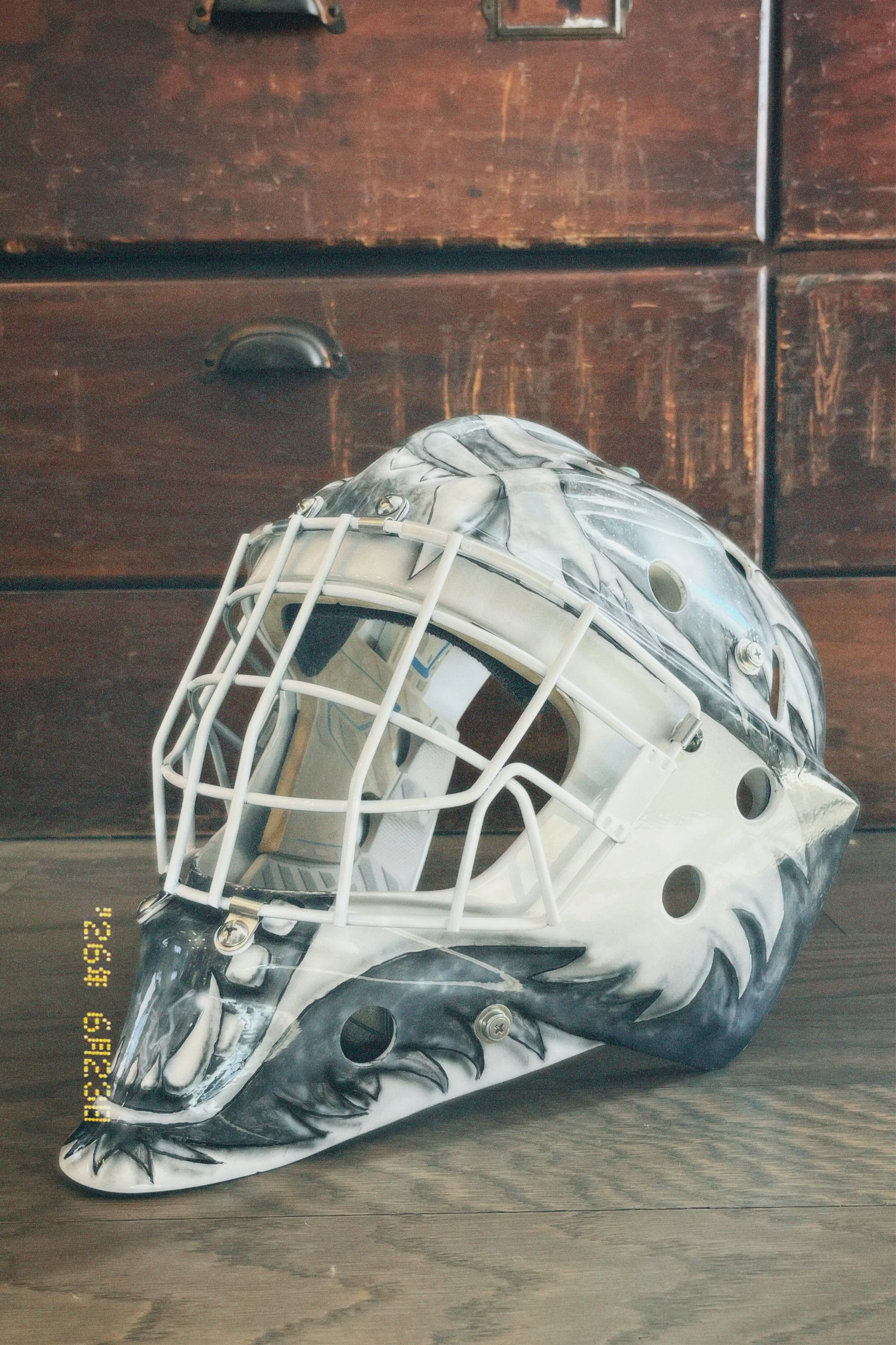 White hockey goalie mask with black and gray tiger face design on a dark wooden surface.