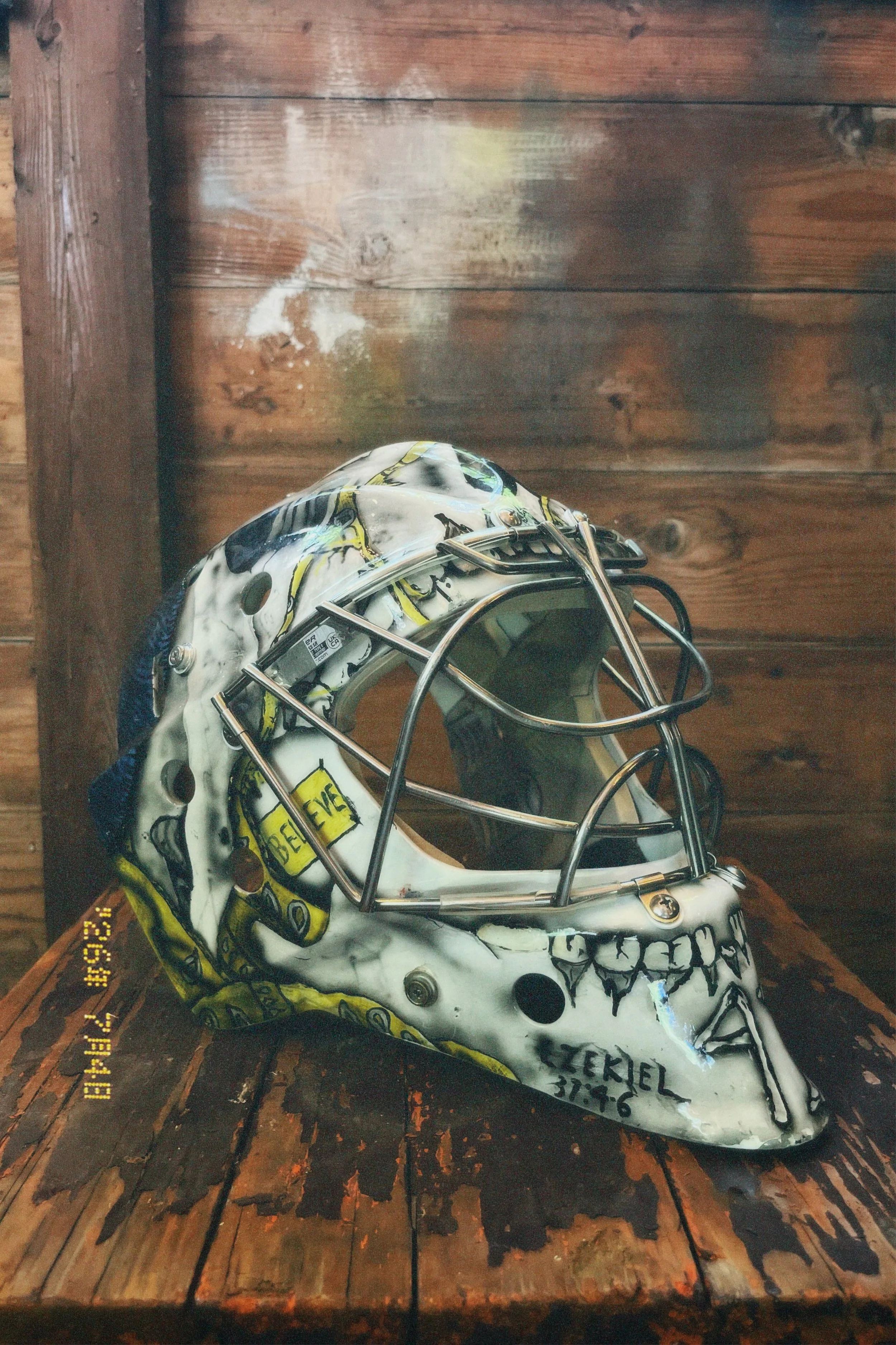 A hockey mask decorated with a skeleton face design, placed on a wooden surface against a wooden wall background.