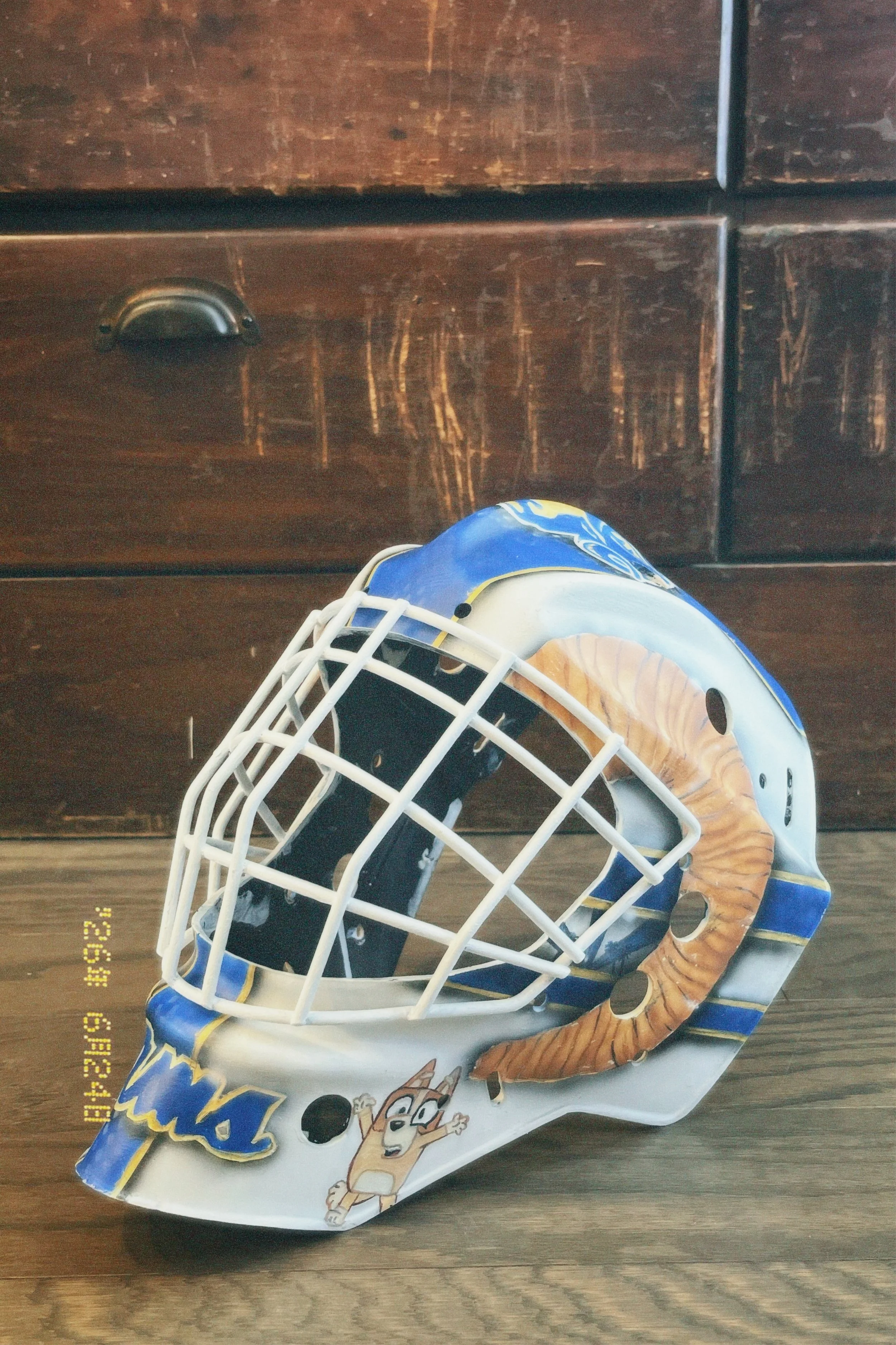Ice hockey goalie mask with Detroit Lions logo and cartoon character, placed on wooden floor in front of a dark wooden dresser.