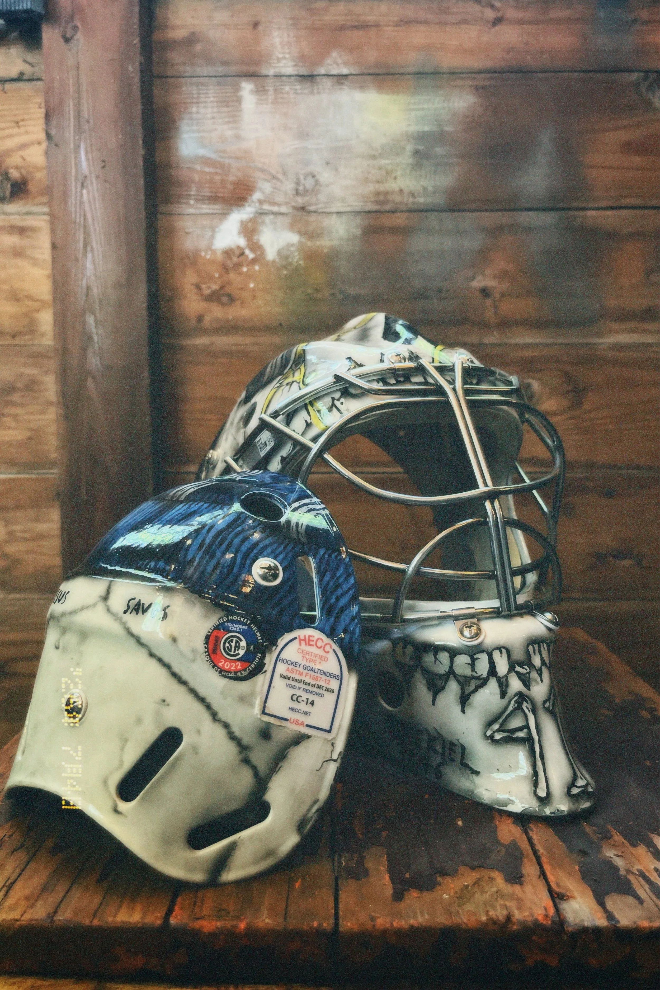 A white ice hockey goalie helmet with black and blue design elements, situated on a wooden surface with a brown wooden wall in the background.